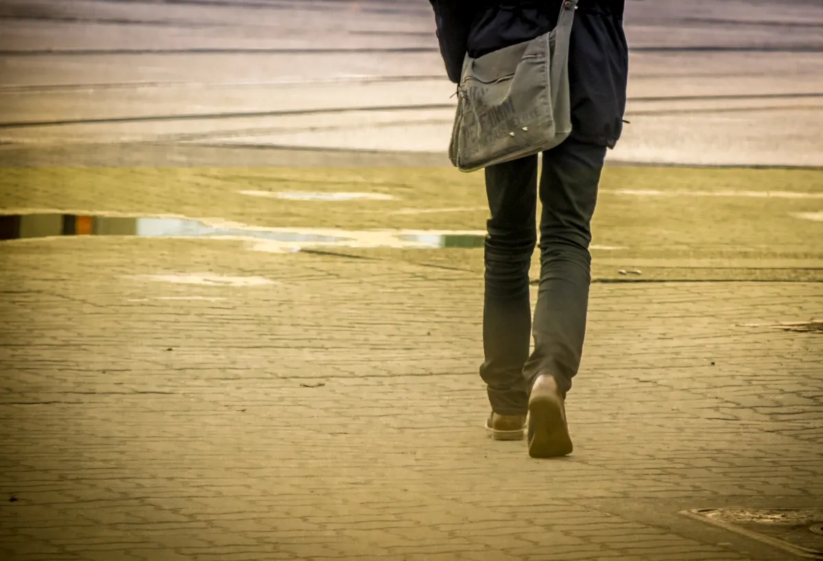 Close-up of a person walking away with a shoulder bag across wet yellow-toned pavement
